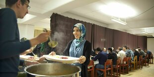 Eine Frau mit Kopftuch lässt sich ihren Teller aus einem großen Topf mit Essen befüllen. Andere Menschen sitzen an Tischen im Hintergrund