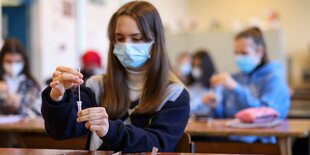 Eine junge Frau sitzt mit Maske an einer Bank in einem Klassenraum. Sie hält ein Wattestäbchen in der einen und eine Lösung in einem kleinen Gefäß in der anderen Hand