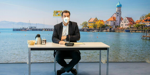 Markus Söders sitz mit Maske an einem Tisch vor dem Hintergrund eines Bayernpanoramas