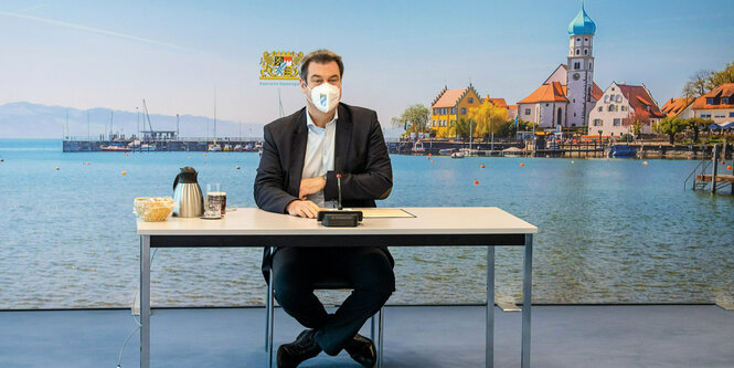 Markus Söders sitz mit Maske an einem Tisch vor dem Hintergrund eines Bayernpanoramas