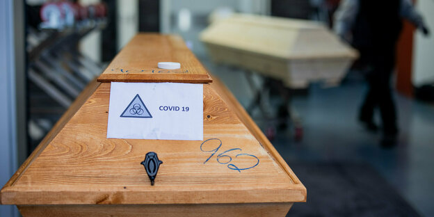 Ein Sarg aus Holz an dem ein Schild befestigt ist auf dem "Covid-19" steht Ein Sarg aus Holz an dem ein Schild befestigt ist auf dem "Covid-19" steht