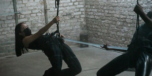 Szene aus Constantin Hartensteins Video "Suspend": Zwei Performer:innen hängen an Tragegurten vor einer Bachsteinwand