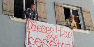 Zwei Männer hängen ein Bettlaken aus dem Fenster eines Hauses mit Fensterläden. Darauf steht: "Dieses Haus ist besetzt"