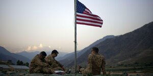 Drei US-Soldaten sitzen vor einer Bergkulisse. Über ihnen weht eine US-Fahne