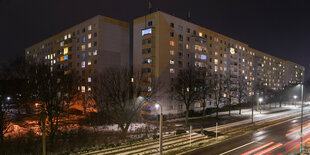 Teilweise hell erleuchtet sind einige Zimmer eines Mehrfamilienhauses am Abend, davor die Gleise einer Tram.