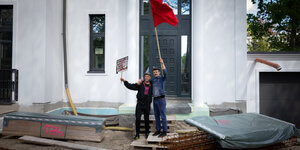 Zwei Männer schwenken vor einem neugebauten Haus eine rote Fahne und tragen ein Schild auf dem steht "Müssen plötzlich ausziehen"