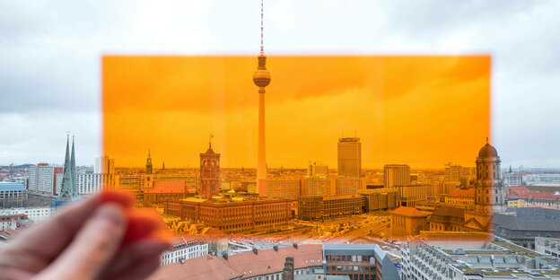 Eine Hand mit orangefarbener tranparenter Schablone vor der grauen Berliner Skyline