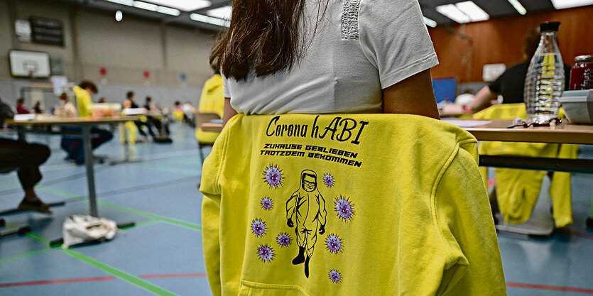 Ein Pullover mit der Aufschrift „Corona hAbi - Zuhause geblieben, trotzdem bekommen“ hängt über der Stuhllehne einer Schülerin, die in einer Turnhalle Abitur schreibt.