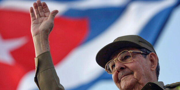 Raul Castro, winkend mit der rechten Hand, unter einer kubanischen Flagge