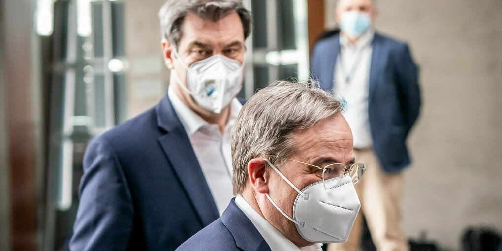 Söder und Laschet stehen versetzt hintereinander und schauen mit ernster Miene in verschiedene Richtungen. Beide tragen FFP2-Masken