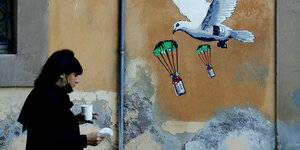 eine Frau läuft mit einem Papp-Becher in Rom an einem Haus vorbei - an der Fassade ist ein Wandbild mit weißer Taube aus derem Bauch Covid-19-Impfstoff abgeworfen wird