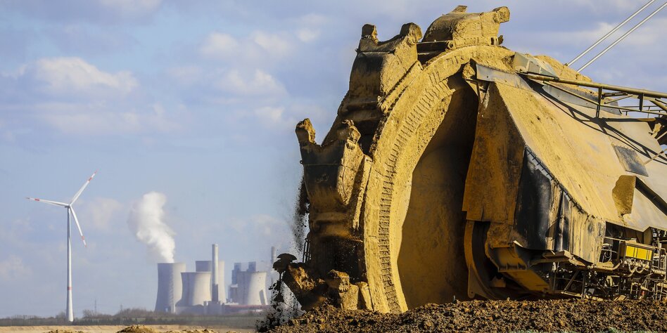 Gelber Schaufelradbagger gräbt Kohle, im Hintegrund Rauchfahne eines Kraftwerks und Windräder