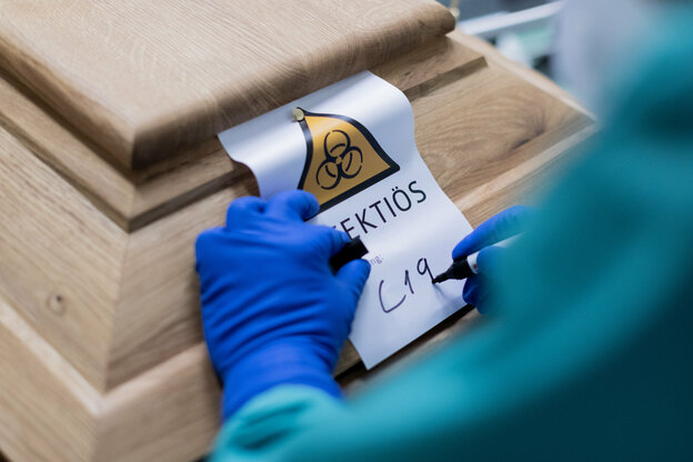 Eine Person in Schutzausrüstung schreibt "C-19" auf einen Zettel, der an einem Sarg befestigt ist Eine Person in Schutzausrüstung schreibt "C-19" auf einen Zettel, der an einem Sarg befestigt ist