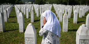 Eine Frau mit Kopftuch und Händen vor dem Gesicht betet auf einem Friedhof mit vielen weißen säulenartigen Grabsteinen mit serbischer und arabischen Inschriften