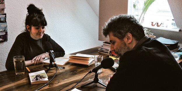 Nefelie Kavouras und Anselm Neft sitzen hinter Mikrophonen an einem Tisch.