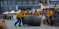 Fünf Männer in gelben T-Shirts rollen ein Weinfass eine Strecke entlang, die mit Strohballen begrenzt ist