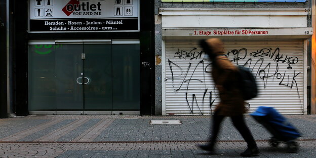 Eine Frau geht mit einem Rollkoffe eine Straße entlang. Im hintergrund sind geschlossene Geschäfte zu sehen