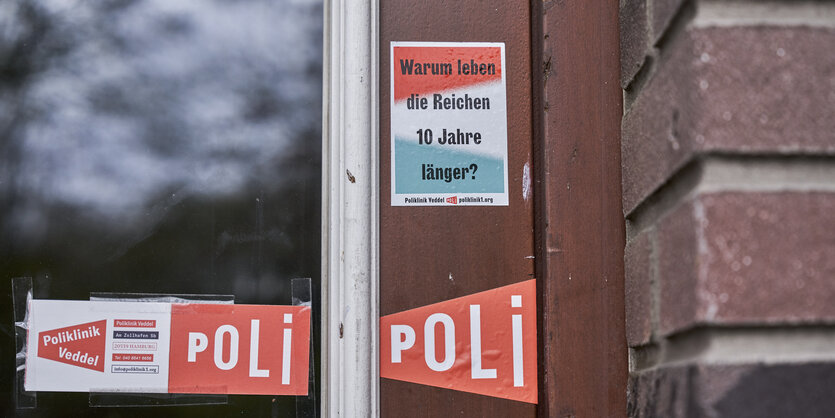 Zwei Aufkleber auf einer Glasscheibe. Auf dem ersten steht "Poliklinik Verddel". Auf dem zweiten steht: "Warum leben die Reichen zehn Jahre länger?"