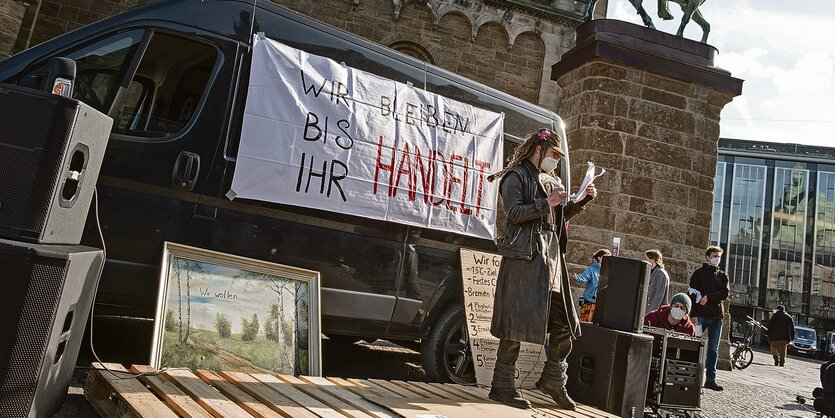 Ein Mann mit Mikrophon steht vor einem Plakat mit der Aufschrift "Wir bleiben bis ihr handelt".