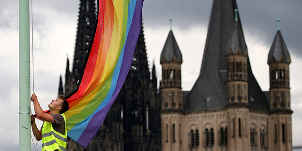 Ein Mann hisst eine Regenbogenflagge vor dem Kölner Dom und der St.Martins Kirche