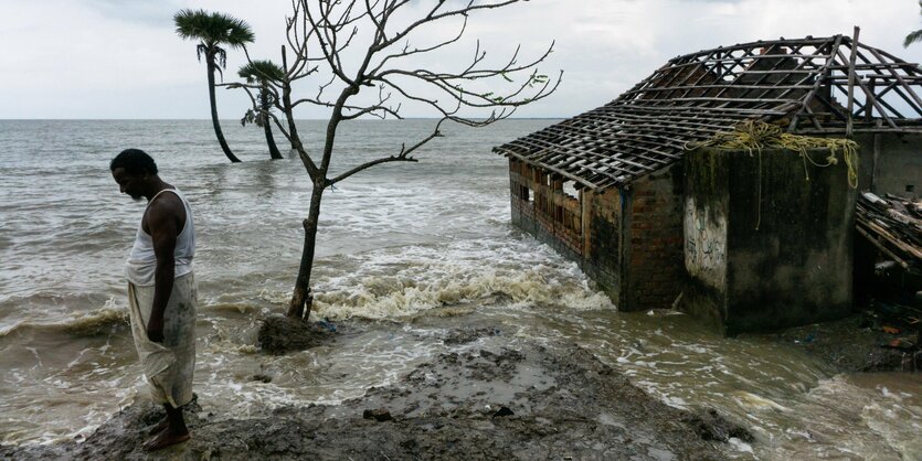 Ein Mann steht an einem überfluteten Strand neben verlassenem Haus - im Wasser zewei vereinzelte Palmen und ein Baum