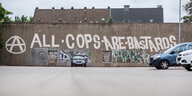 An einer Wand steht "All cops are bastards", davor parken Autos