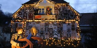 Hänsel und Gretel als Puppen vor dem Lebkuchenhaus. oben aus dem Fenster schaut die Hexe