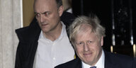 Boris Johnson und Dominic Cummings verlassen ein Gebäude - die Augen von Cummings leuchten durch ein Blitzlich rot