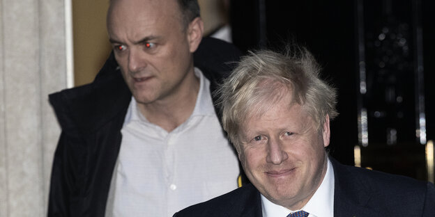 Boris Johnson und Dominic Cummings verlassen ein Gebäude - die Augen von Cummings leuchten durch ein Blitzlich rot