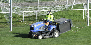 Ein Gemeindemitarbeiter mit Mundschutz sitzt auf einem Rasenmäher und fährt über einen Fußballrasen.