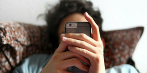 Junger Mann an ein Kissen gelehnt, das Smartphone in der Hand verdeckt sein Gesicht