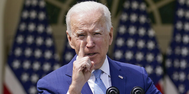 Joe Biden vor US-Flaggen hebt den zeigefinger der rechten Hand