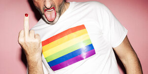 Mann mit Regenbogenflagge auf dem T-Shirt, rotem Nagellack und Lippenstift, zeigt den Stinkefinger