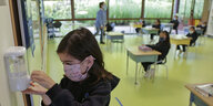 Ein Kind desinfiziert sich die Hände in einem Klassenraum dessen Schultische weit entfernt von einander stehen