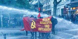 Demonstranten ducken sich hinter einem roten Banner mit der Aufschrift "Gegen Ausbeutung und Unterdrückung" vor den Strahlen der Wasserwerfer.