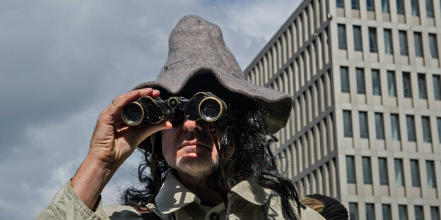 Ein als Spion verkleideter Mann steht mit Fernglas und Schlapphut vor einem Teil des Neubaus des Bundesnachrichtendienstes in Berlin.