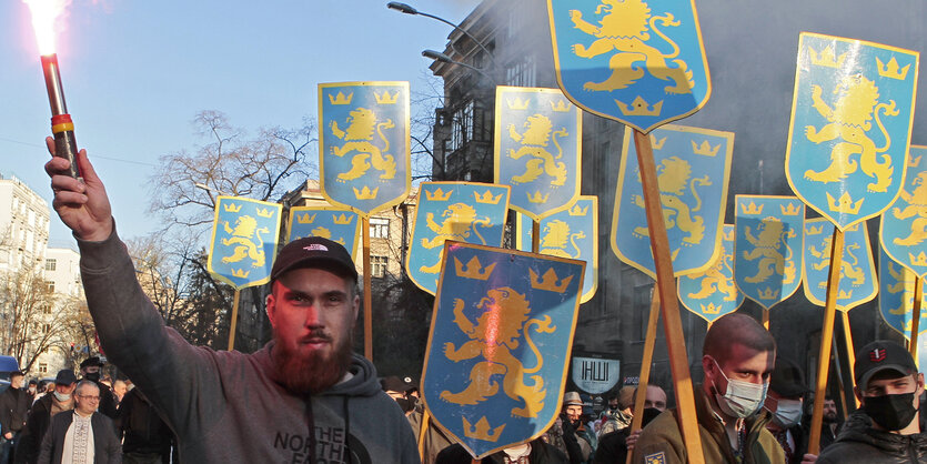 Männer marschieren mit Schildern auf denen ein blau-gelbes Wappen zu sehenist - ein Mann hält ein bengalisches feuer in der Hand