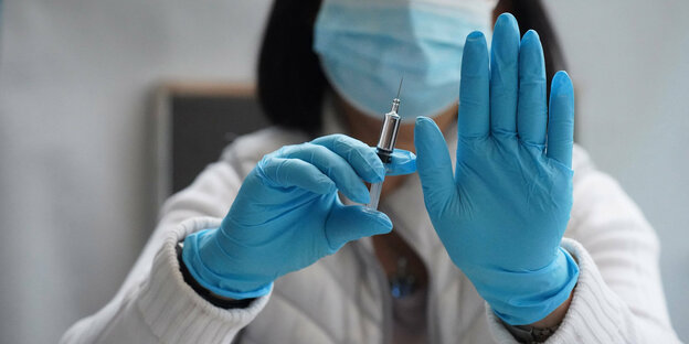 Eine Ärztin mit Mundschutz und Schutzhandschuhen hält eine Spritze in der einen Hand und macht eine ablehnende Geste mit der anderen Hand
