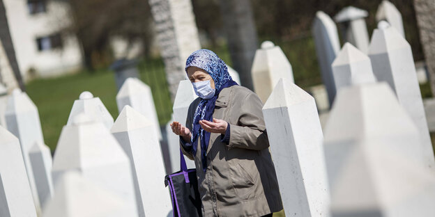 Eine alte Frau mit Kopftuch und Mundschutz betet auf einem Friedhof mit weißen säulenförmigen Grabsteinen