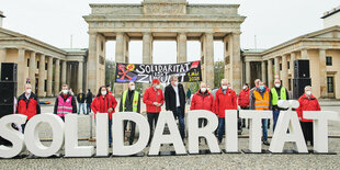 Große Buchstaben bilden das Wort Solidarität, dahinter stehen Menschen mit Mund-Nasen-Schutz. Das Ganze spielt sich auf dem Pariser Platz vor dem Brandenurger Tor ab.