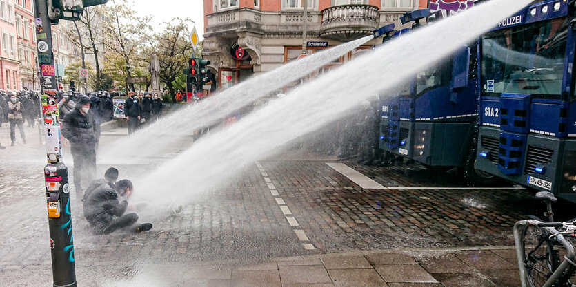 2 Personen sitzen auf der Straße, ein Wasserwerfer schießt seinen Wasserstrahl gezielt auf sie