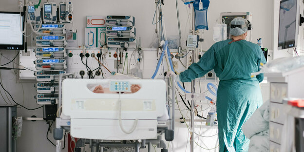 Eine Person in Schutzkleidung steht neben einem Bett an das zahlreiche medizinische Geräte angeschlossen sind Eine Person in Schutzkleidung steht neben einem Bett an das zahlreiche medizinische Geräte angeschlossen sind