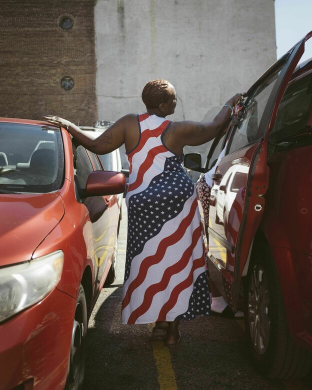 Eine Frau mit Kleid in US-Flaggen-Muster steht zwischen zwei Autos.