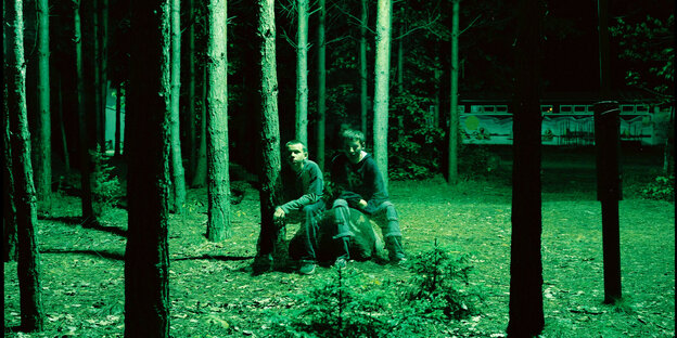 In einem Wald, grünes Licht, es wirkt dunkel, zwei Jungen sitzen auf einem Baumstumpf
