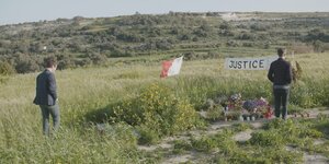 Zwei Männer stehen auf einer Wiese in Malta vor Grabkerzen und einer Fahne mit der Aufschrift "Justice".