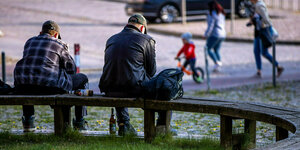 Zwei junge Männer spielen mit ihren Mobiltelefonen auf einer runden Bank am Marktplatz und trinken Bier.