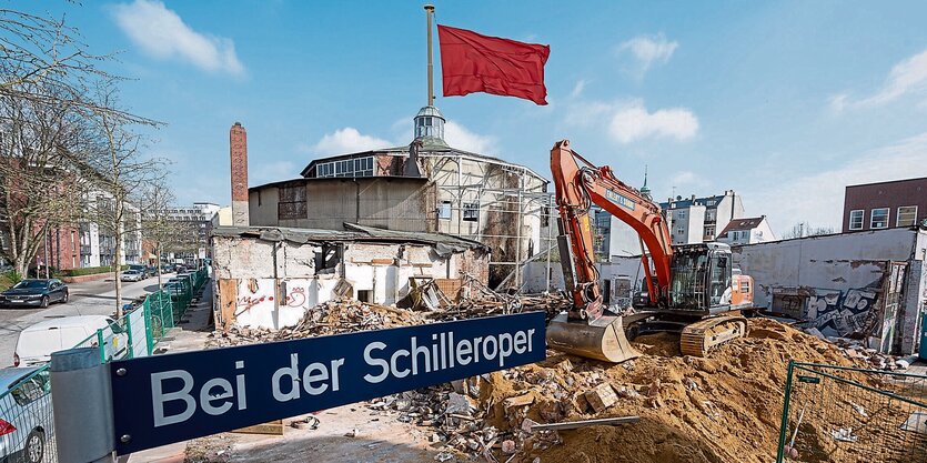 Eine rote Fahne weht auf einem Rundbau, davor halb abgerissene Gebäude, ein Bagger und ein Straßenschild