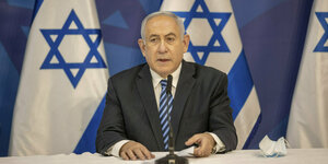 Benjamin Netanjahu sitzt vor israelischen Flaggen hinter einem Mikrophon