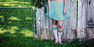 Eine Person mit Regenbogen-Socken und grünem Rock steht vor einem Zaun.