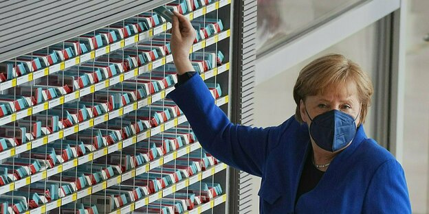 Angela Merkel zieht eine Stimmkarte im Bundestag. Sie trägt eien blaue Maske.